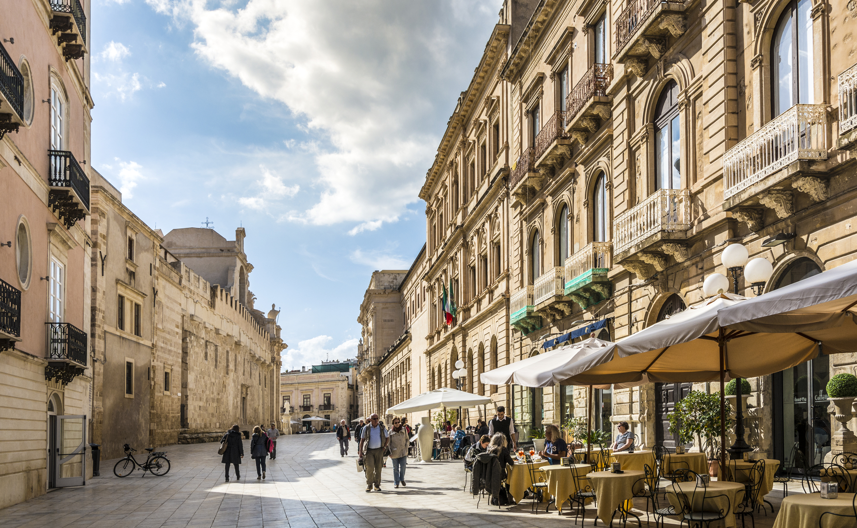 Syracuse, Italy - March 17, 2016: People of various ages walking and sitting in the centre of Syracuse (Siracusa in Italian), a major town and tourist destination in Sicily, Italy. There are cafes on the right and the Cathedral on the left. Daytime.