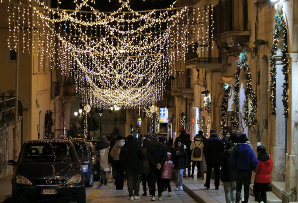 A crowd walks down a high street with clusters of festive lights over head