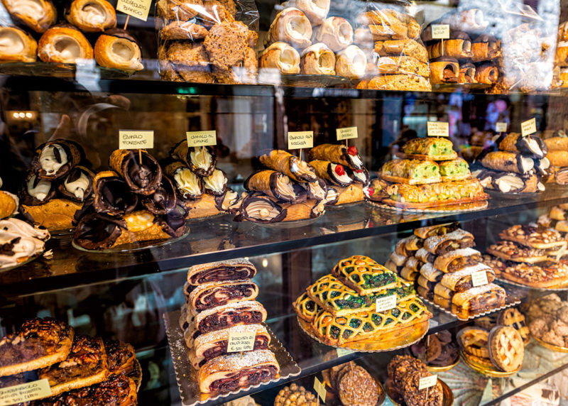 Pastry bakery store shop in Assisi, Umbria Italy selling dessert chocolate cannoli, rocciata cakes and pies with price tags through retail display glass window